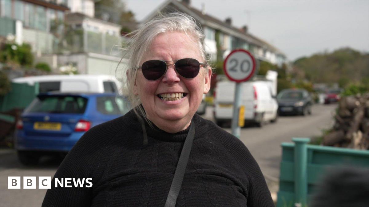 A woman standing on a street. She is wearing sunglasses and is smiling into the camera. Parked cars and houses are behind her.