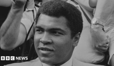 A black and white image of Muhammad Ali smiling and looking to the left of the camera. He is wearing a suit and behind him is a photographer holding a camera.