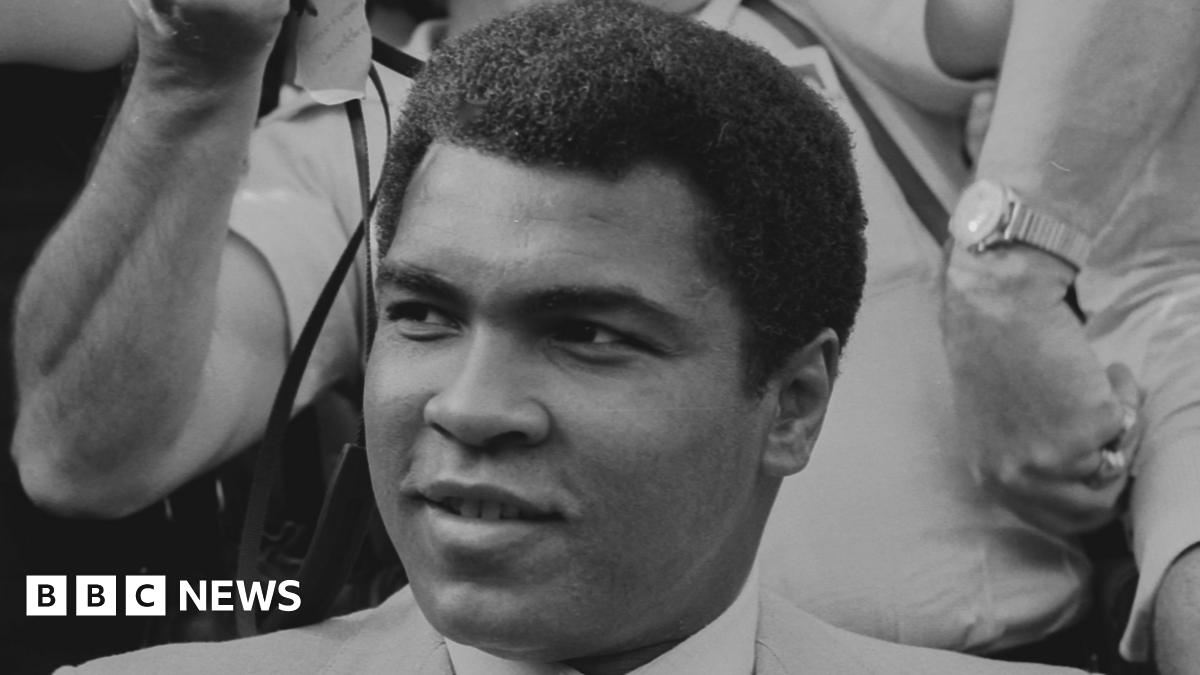 A black and white image of Muhammad Ali smiling and looking to the left of the camera. He is wearing a suit and behind him is a photographer holding a camera.