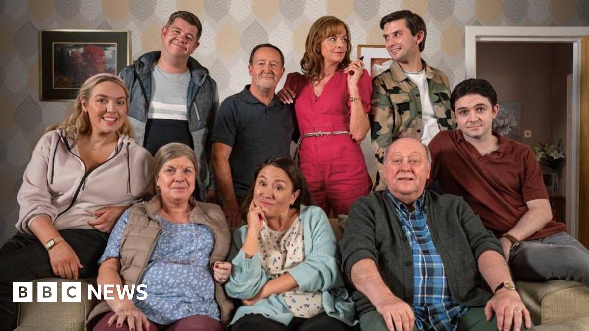 A group of eight people gathered in a living room setting. Four individuals are seated on a couch in the foreground, while four others stand behind them. The background features patterned wallpaper with a leaf design in muted tones.