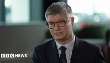 Lord Wolfson, chief executive of Next. He is sitting in a chair during an interview with the BBC. He is wearing glasses, a spotted tie, and a dark blue suit.