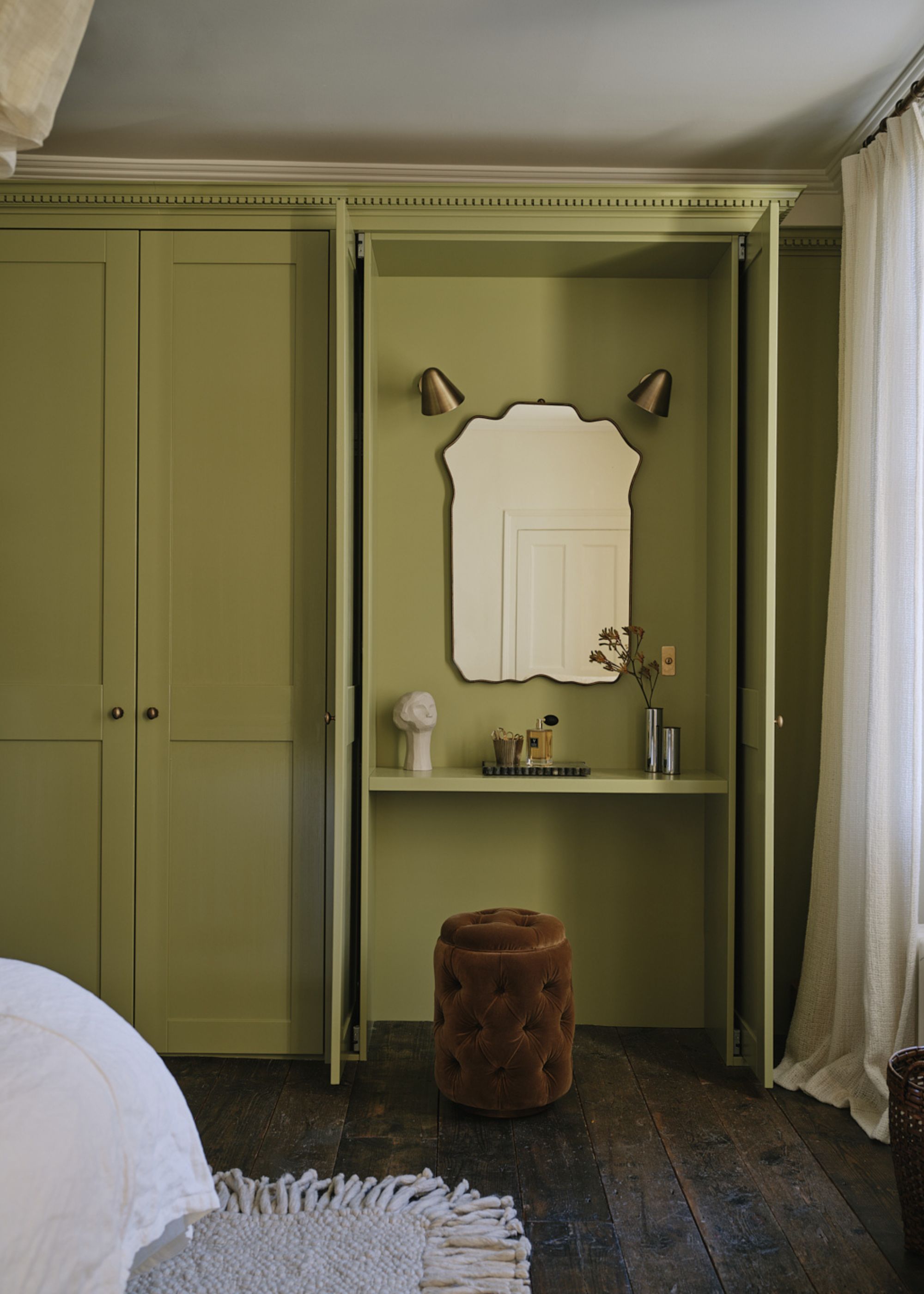 A bedroom with built-in wardrobes, color-drenched in a warm shade of green. Original dark wooden floorboards, sheer curtains at the window, and a vanity setup inside the wardrobe.