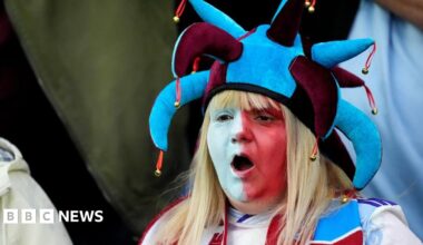 An Aston Villa fan in the stands before the UEFA Europa League match at Villa Park, Birmingham. She has long blonde hair and her face is painted half white, half red. She is wearing a maroon and blue jesters hat and her mouth is open as if mid chant or song.