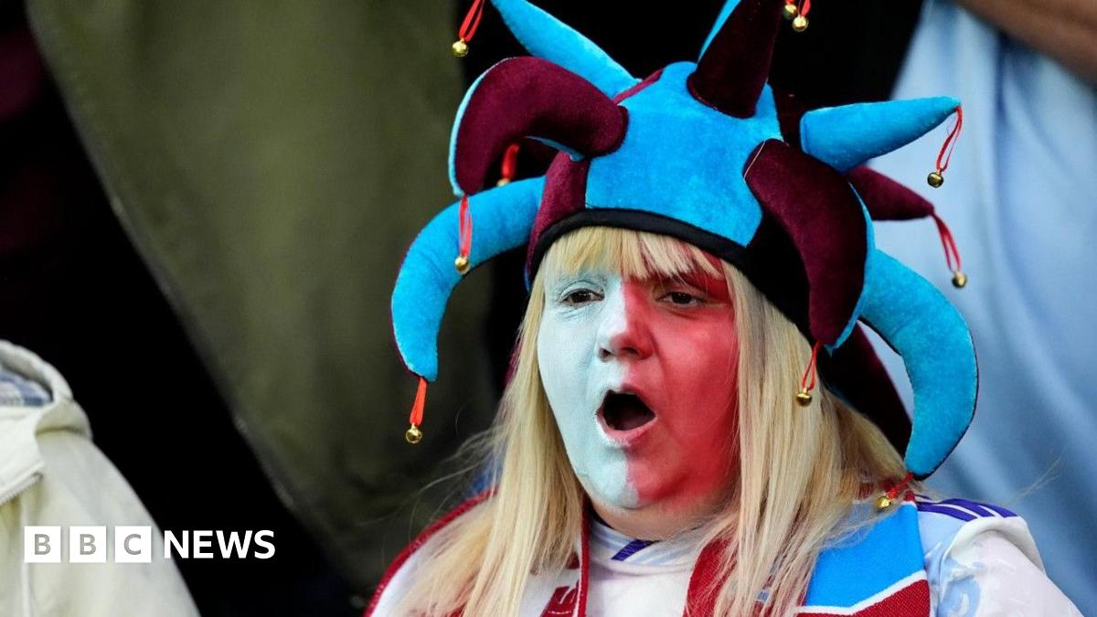 An Aston Villa fan in the stands before the UEFA Europa League match at Villa Park, Birmingham. She has long blonde hair and her face is painted half white, half red. She is wearing a maroon and blue jesters hat and her mouth is open as if mid chant or song.