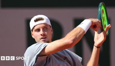 British tennis player Toby Samuel playing a forehand during a match at the French Open in Paris. Samuel is wearing a grey shirt and white hat, with the visor back to front.