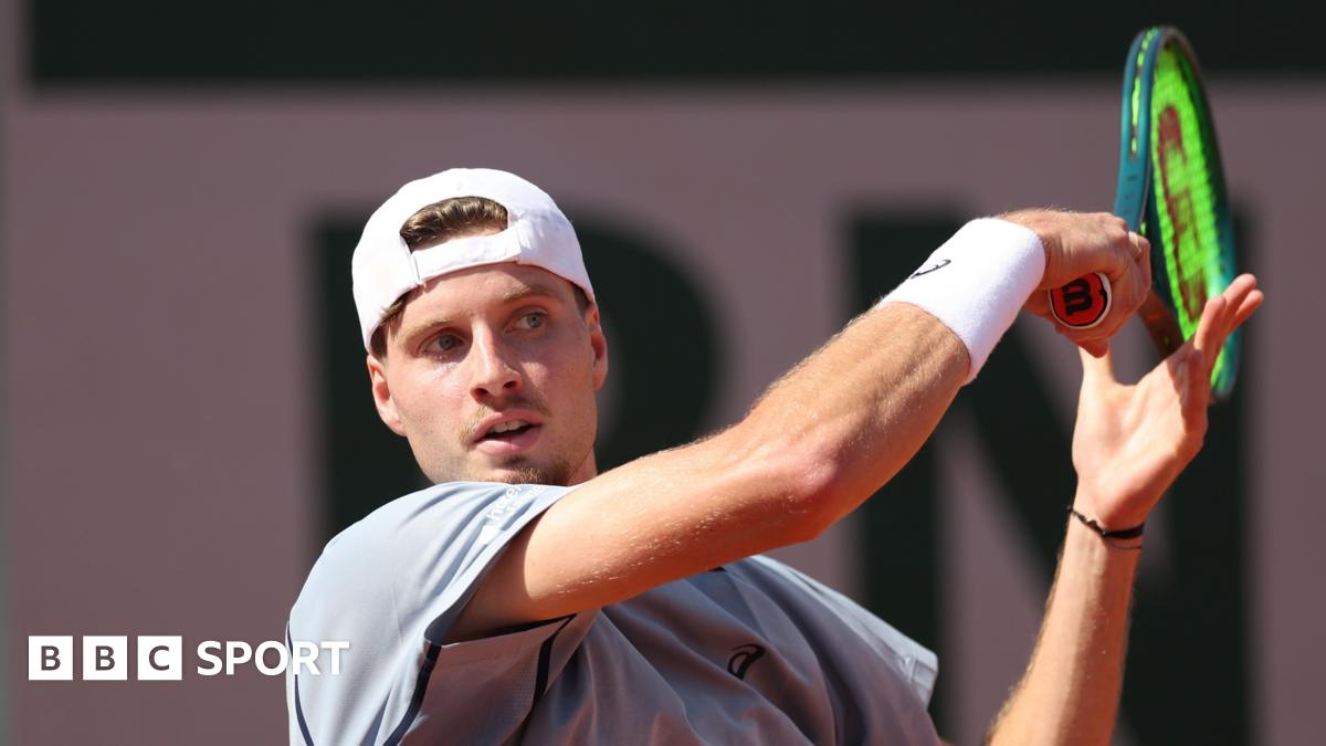 British tennis player Toby Samuel playing a forehand during a match at the French Open in Paris. Samuel is wearing a grey shirt and white hat, with the visor back to front.