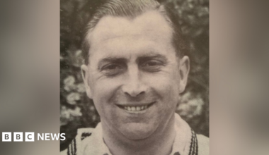 Black and white portrait of Harold Gimblett wearing a white shirt and sweater and smiling.