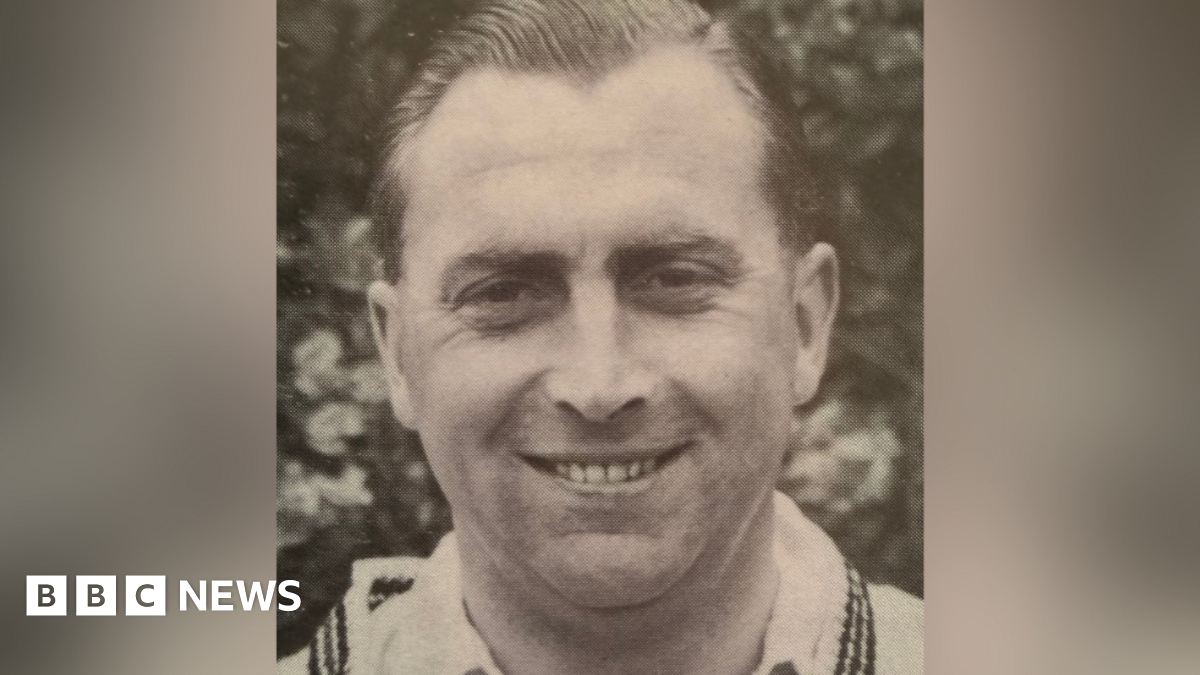 Black and white portrait of Harold Gimblett wearing a white shirt and sweater and smiling.
