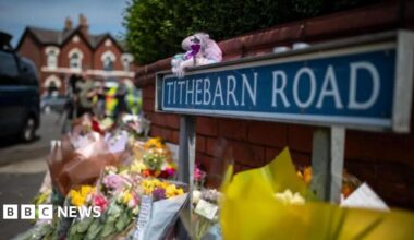 Bunches of flowers and teddies lined up against a wall next to a road sign reading Tithebarn Road.