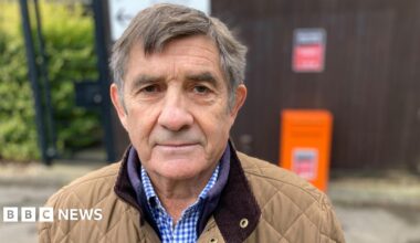 Philip is wearing a brown quilted jacket and a gingham blue shirt. He has short brown and grey hair and has brown eyes. He is looking into the camera.