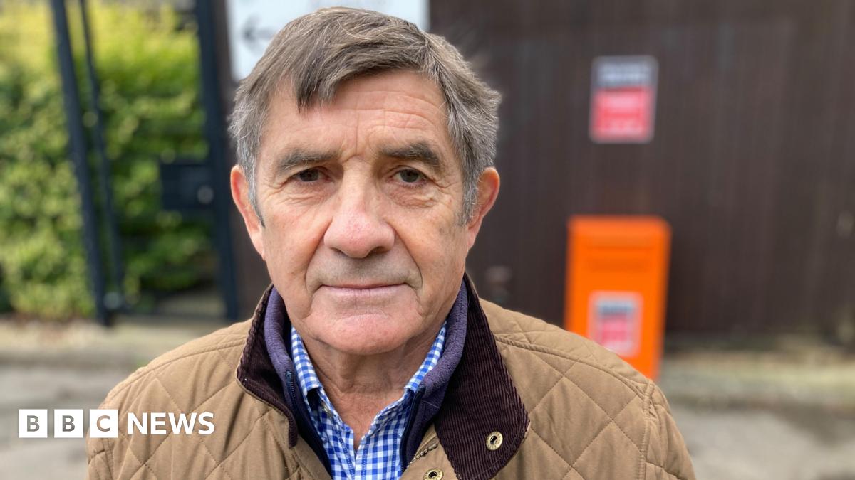 Philip is wearing a brown quilted jacket and a gingham blue shirt. He has short brown and grey hair and has brown eyes. He is looking into the camera.