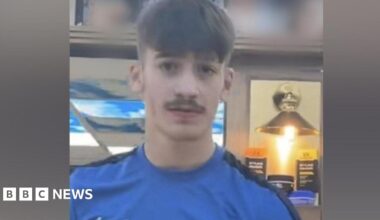 A picture of Taylor wearing a blue shirt with a white Nike logo on it. He has a short moustache and short hair.