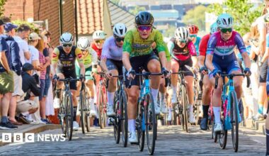 A group of cyclists wearing colourful cycling kit riding towards the camera on a narrow cobbled street. Their faces show the pain and effort of climbing up a steep hill. Spectators line the paths next to the road, with a view looking downhill towards the buildings of a city centre in the background.