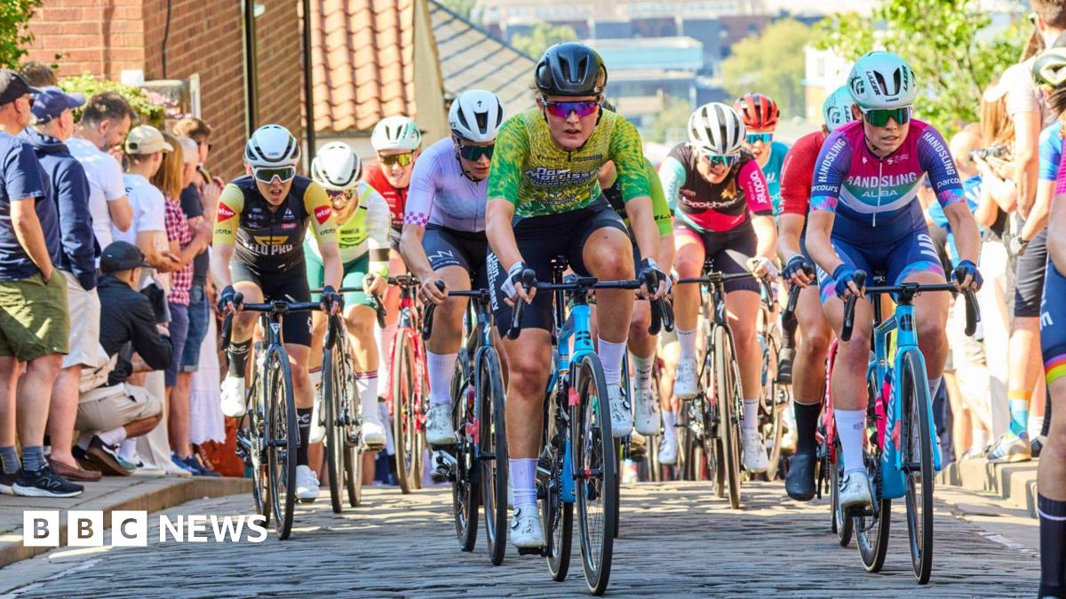 A group of cyclists wearing colourful cycling kit riding towards the camera on a narrow cobbled street. Their faces show the pain and effort of climbing up a steep hill. Spectators line the paths next to the road, with a view looking downhill towards the buildings of a city centre in the background.