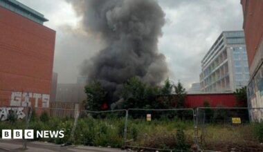 Smoke billowing from a fire, in a huge plume going upward, behind a wall which is behind a metal fence.