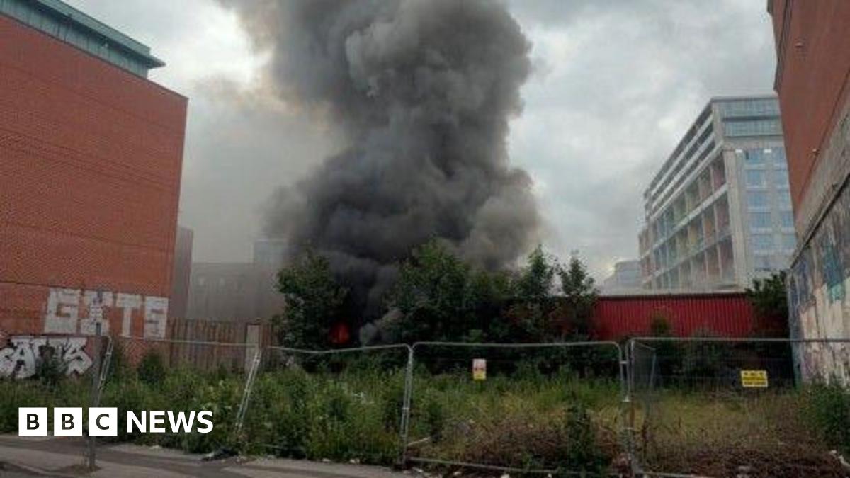 Smoke billowing from a fire, in a huge plume going upward, behind a wall which is behind a metal fence.