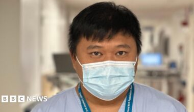 Tito Vicario is an Asian nurse, wearing blue scrubs and a lanyard. He is also wearing a blue mask. He has black straight hair and dark eyes. He's standing on his ward and in the background are blurred-out computers and equipment.