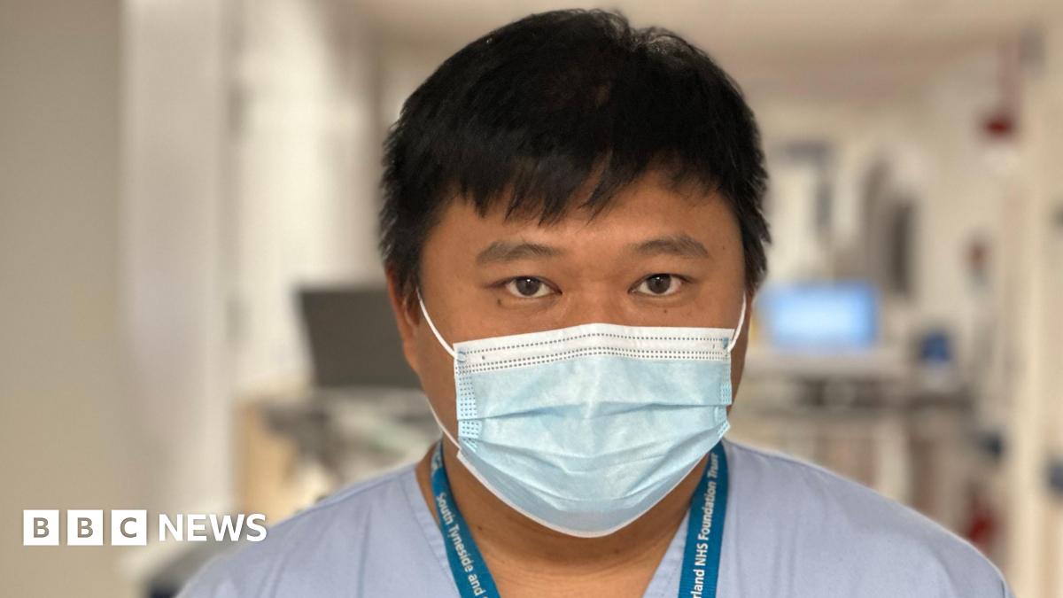 Tito Vicario is an Asian nurse, wearing blue scrubs and a lanyard. He is also wearing a blue mask. He has black straight hair and dark eyes. He's standing on his ward and in the background are blurred-out computers and equipment.