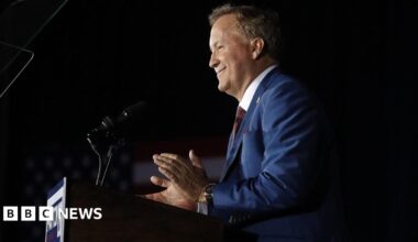 Attorney General Ken Paxton is seen in profile at a podium with a dark background from the waist up. He appears to be smiling and clapping and his campaign sign is on the front of the podium.