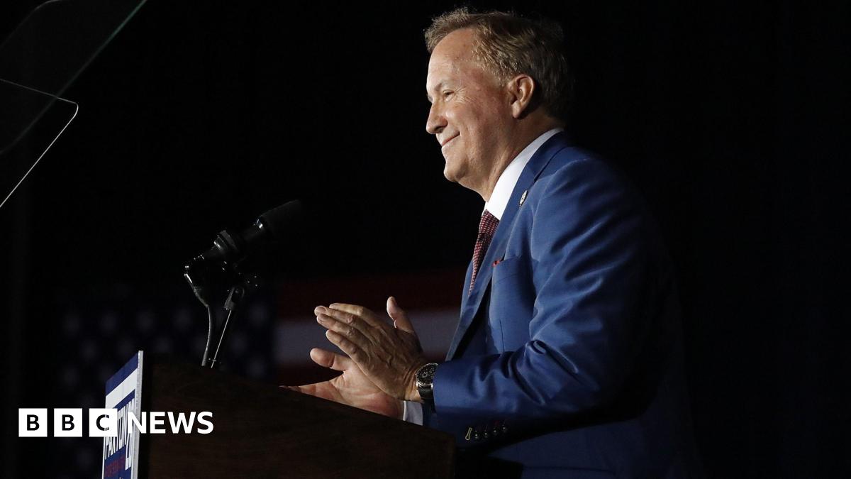 Attorney General Ken Paxton is seen in profile at a podium with a dark background from the waist up. He appears to be smiling and clapping and his campaign sign is on the front of the podium.