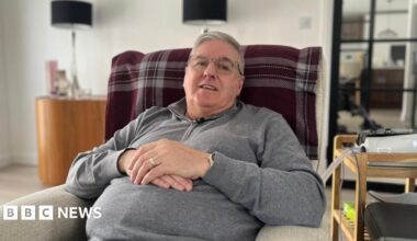 Steven Barrett sits in an armchair in a living room, facing the camera and smiling. He is wearing a grey fleece and glasses.