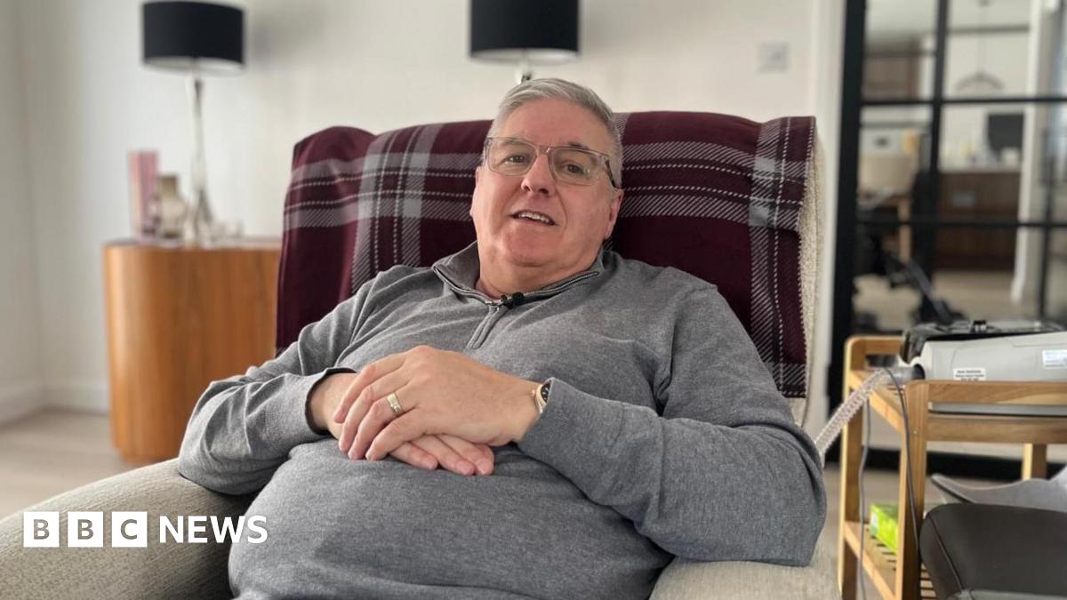 Steven Barrett sits in an armchair in a living room, facing the camera and smiling. He is wearing a grey fleece and glasses.