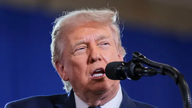 Close up of US President Donald Trump mid-speech. He's in a dark suit, white shirt and tie, speaking into thin black mics