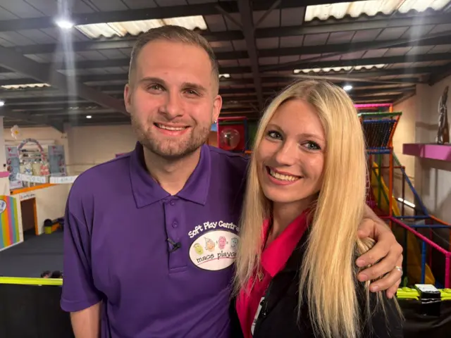 Boston wearing a purple soft play centre branded polo shirt and smiling with his arm around his sister's shoulders. She has long straight blonde hair and is also smiling wearing a red polo shirt with the same branding and a black cardigan