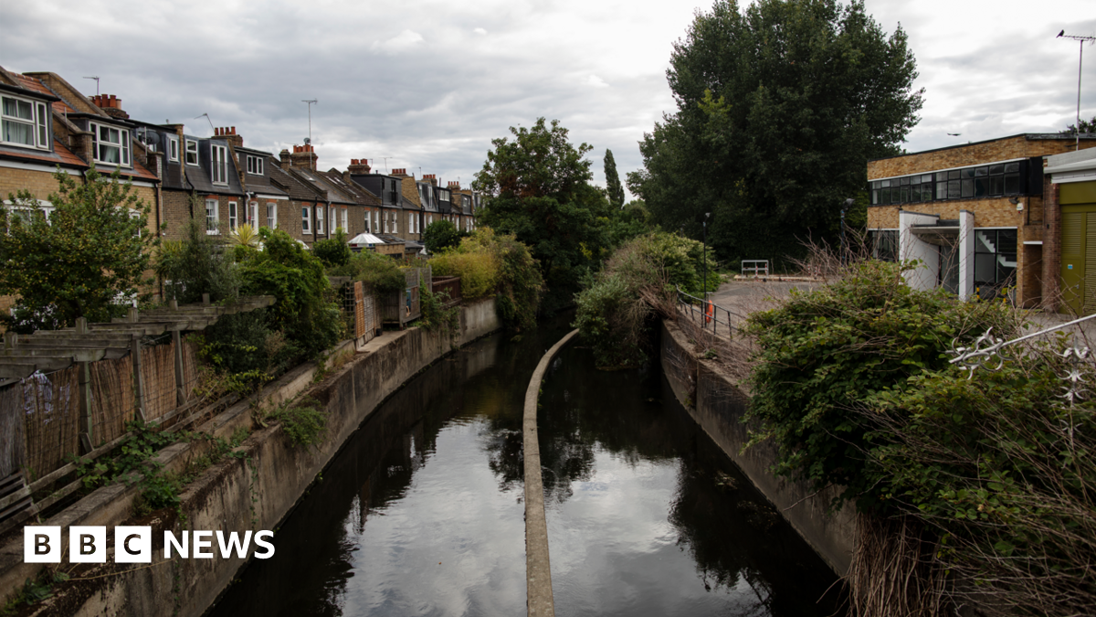 The river Wandle runs past houses in Wandsworth on August 24, 2020 in London, England. The river is in the middle and is enclosed by concrete banks on either side. On the left is a row of terraced houses and on the right is a modern brick and glass 1960s style building. There is some vegetation on the river banks.