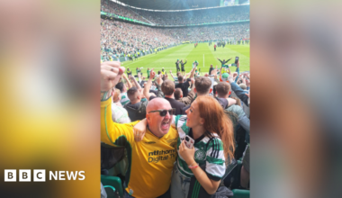 A man wearing a yellow Celtic jersey is smiling and raising his fist in the air. He has his arm around a young woman who is also smiling and wearing a green and white Celtic jersey. They are standing in a football stadium and behind them lot of fans are celebrating.