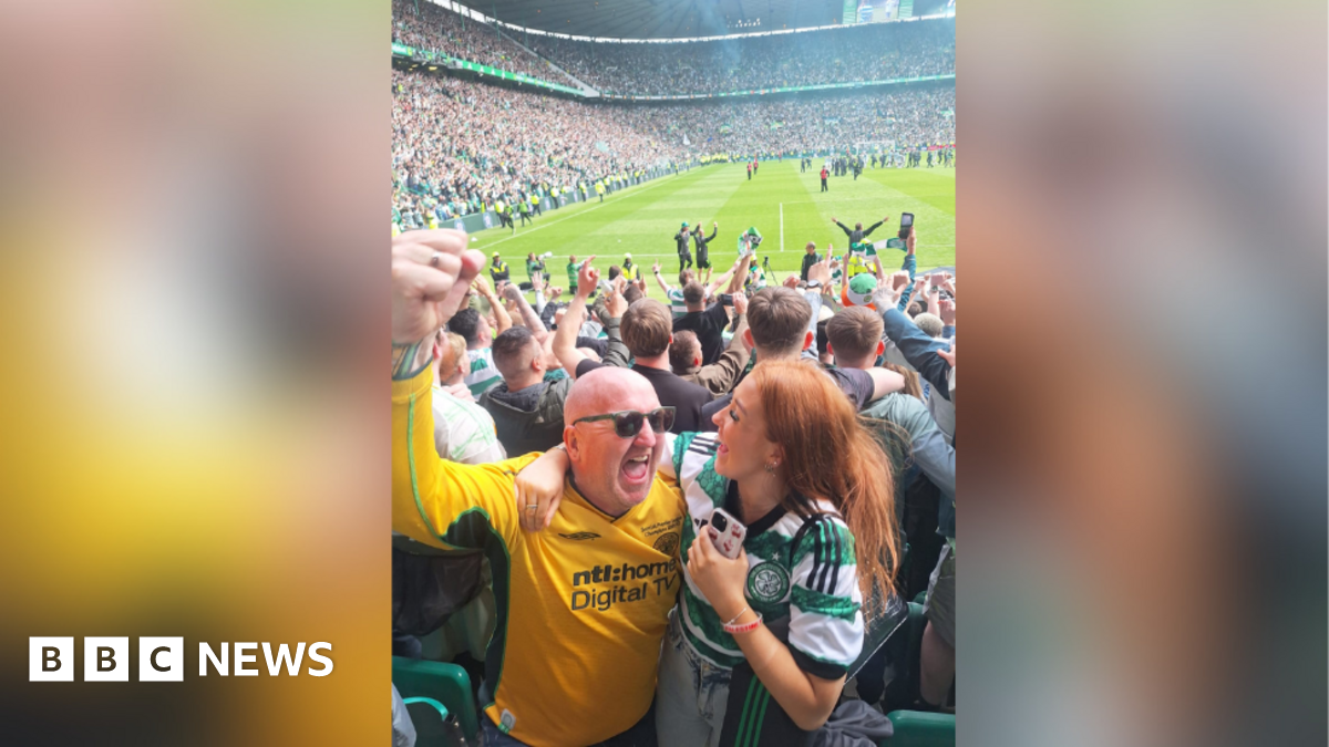 A man wearing a yellow Celtic jersey is smiling and raising his fist in the air. He has his arm around a young woman who is also smiling and wearing a green and white Celtic jersey. They are standing in a football stadium and behind them lot of fans are celebrating.