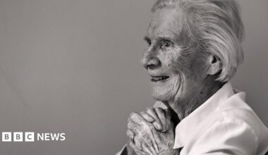 A black and white photo of an elderly lady sitting in a chair against a plain wall, smiling with her hands clasped as she looks out of the frame.