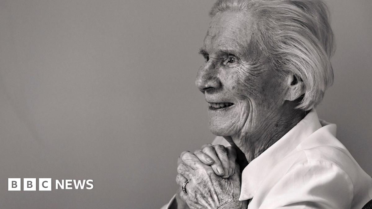 A black and white photo of an elderly lady sitting in a chair against a plain wall, smiling with her hands clasped as she looks out of the frame.