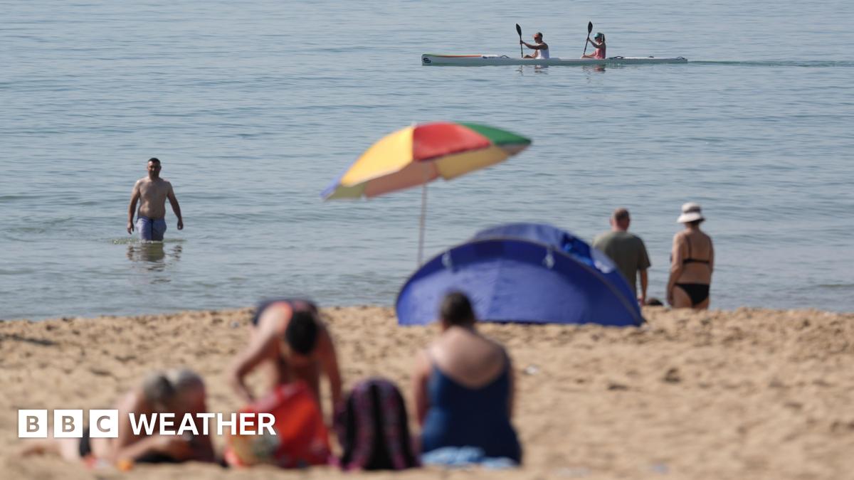 People on beach during May heatwave. One man wades in the sea whist two people paddle by in a kayak