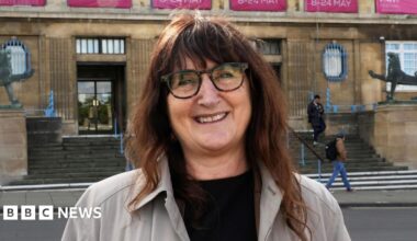 Lucy Galvin, Green leader of Norwich City Council standing outside of City Hall. She is looking directly at the camera and is smiling. She is wearing a black T-shirt with a grey jacket over the top and has brunette hair and a pair of brown framed glasses over her eyes.