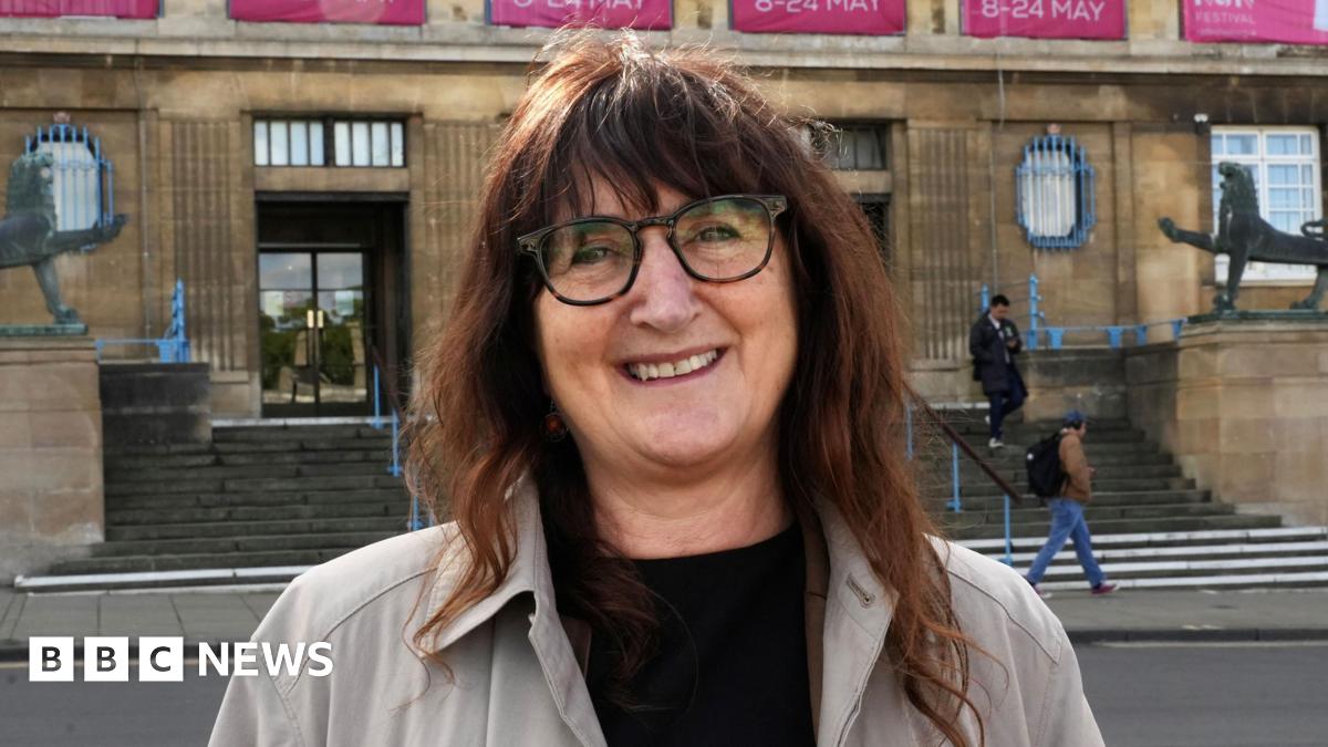 Lucy Galvin, Green leader of Norwich City Council standing outside of City Hall. She is looking directly at the camera and is smiling. She is wearing a black T-shirt with a grey jacket over the top and has brunette hair and a pair of brown framed glasses over her eyes.