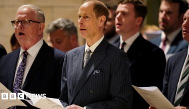 MSPs and guests stand holding service programmes during the Royal Kirking of Parliament at St Giles' Cathedral in Edinburgh. The group, dressed in formal attire, faces the front of the cathedral during the ceremonial church service beneath the building’s stone arches and warm lighting.