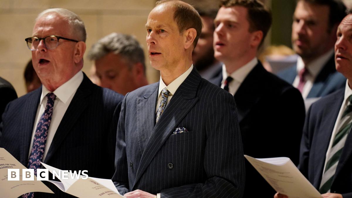 MSPs and guests stand holding service programmes during the Royal Kirking of Parliament at St Giles' Cathedral in Edinburgh. The group, dressed in formal attire, faces the front of the cathedral during the ceremonial church service beneath the building’s stone arches and warm lighting.