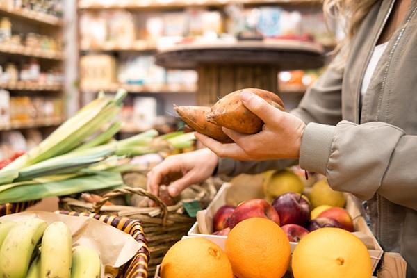 Close-up of the hands of a woman choosing yams from a basket at a market; other kinds of vegetables are arranged on the table in front of her.