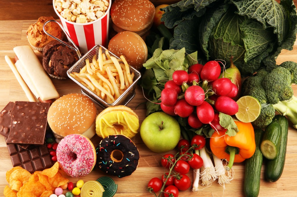 A dichotomy of healthy and unhealthy foods on a wooden table, with fresh produce on the right and junk food on the left.