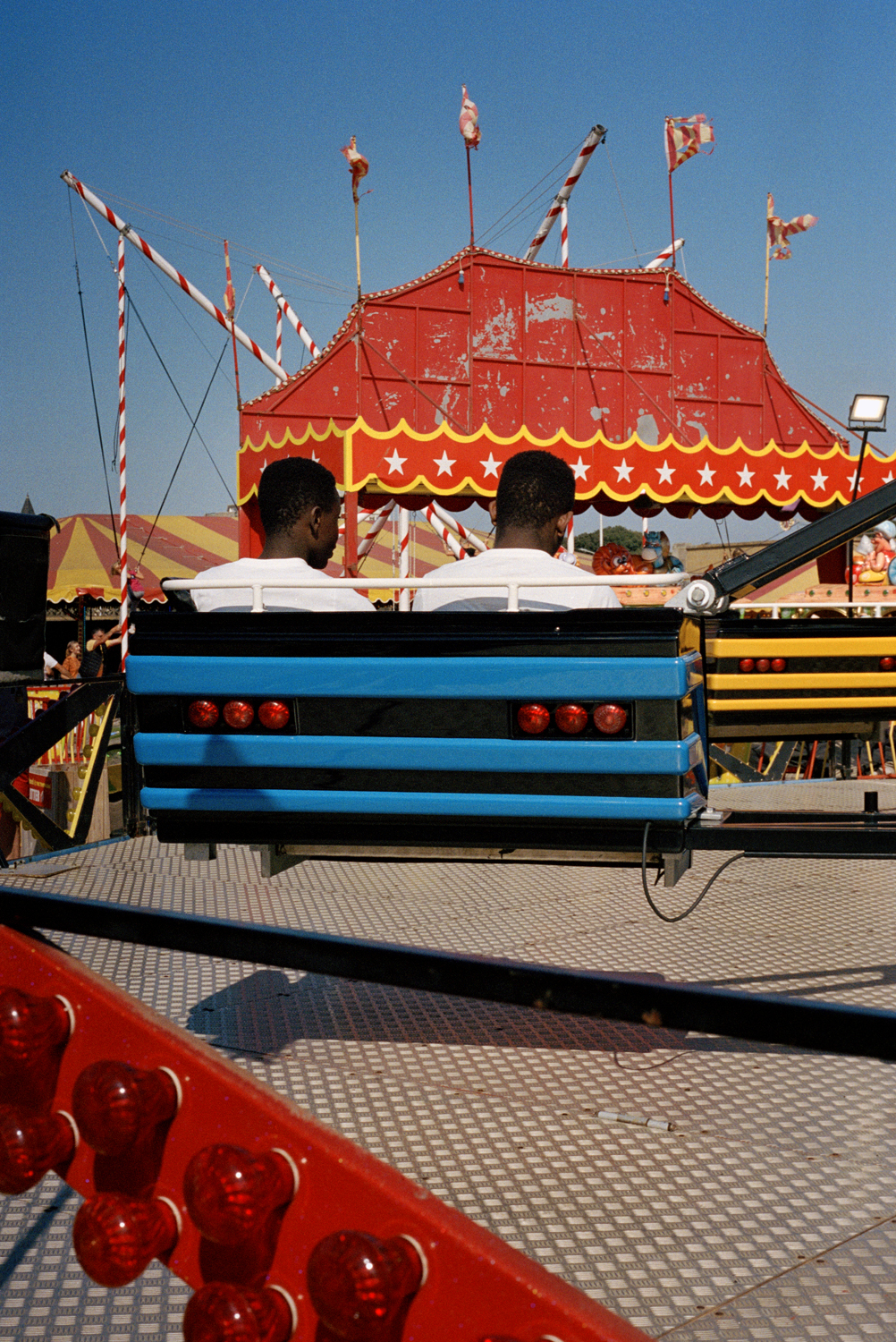 Two young men in matching white t-shirts sit back-to-back in a fairground ride car, a peeling red and gold circus tent rising behind them against a clear blue sky at Weston-Super-Mare.