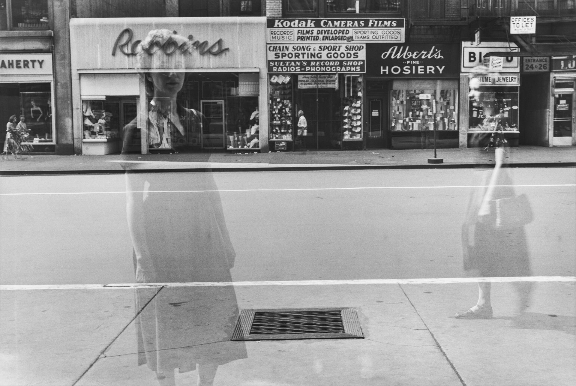 A double-exposure layers a ghostly female figure over a New York street scene, her translucent form standing in front of shop fronts including a Kodak Cameras Films sign.