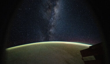 The Milky Way galaxy appears above Earth's greenish atmospheric glow. The galaxy's dust and stars can be seen against the darkness of space.