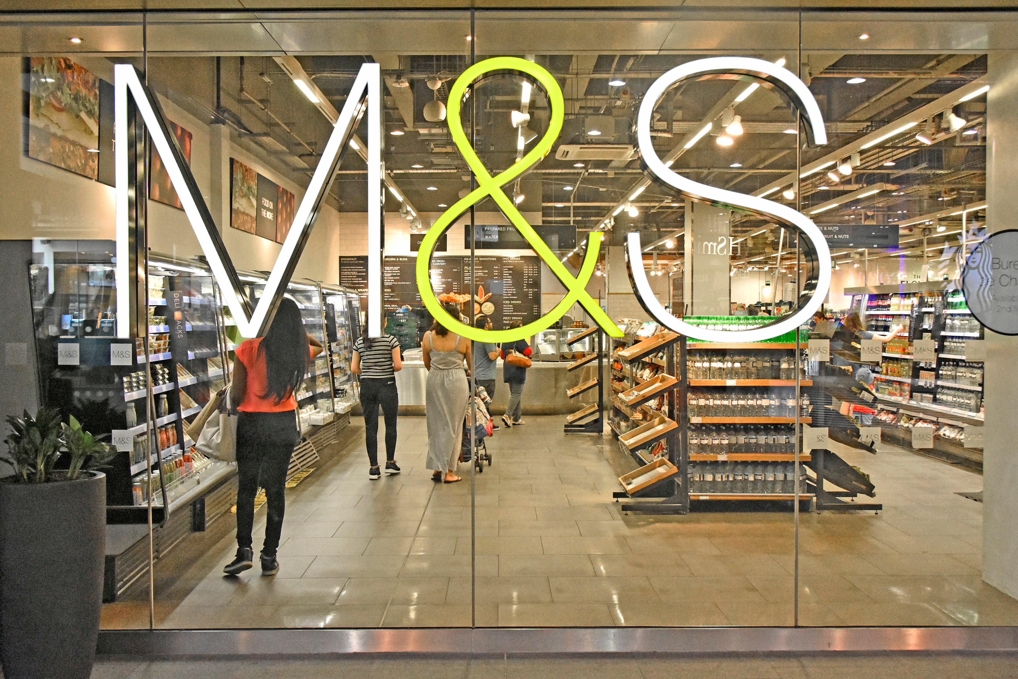 M&S sign on the glass window of a food hall in a Marks and Spencer store, with shoppers inside.