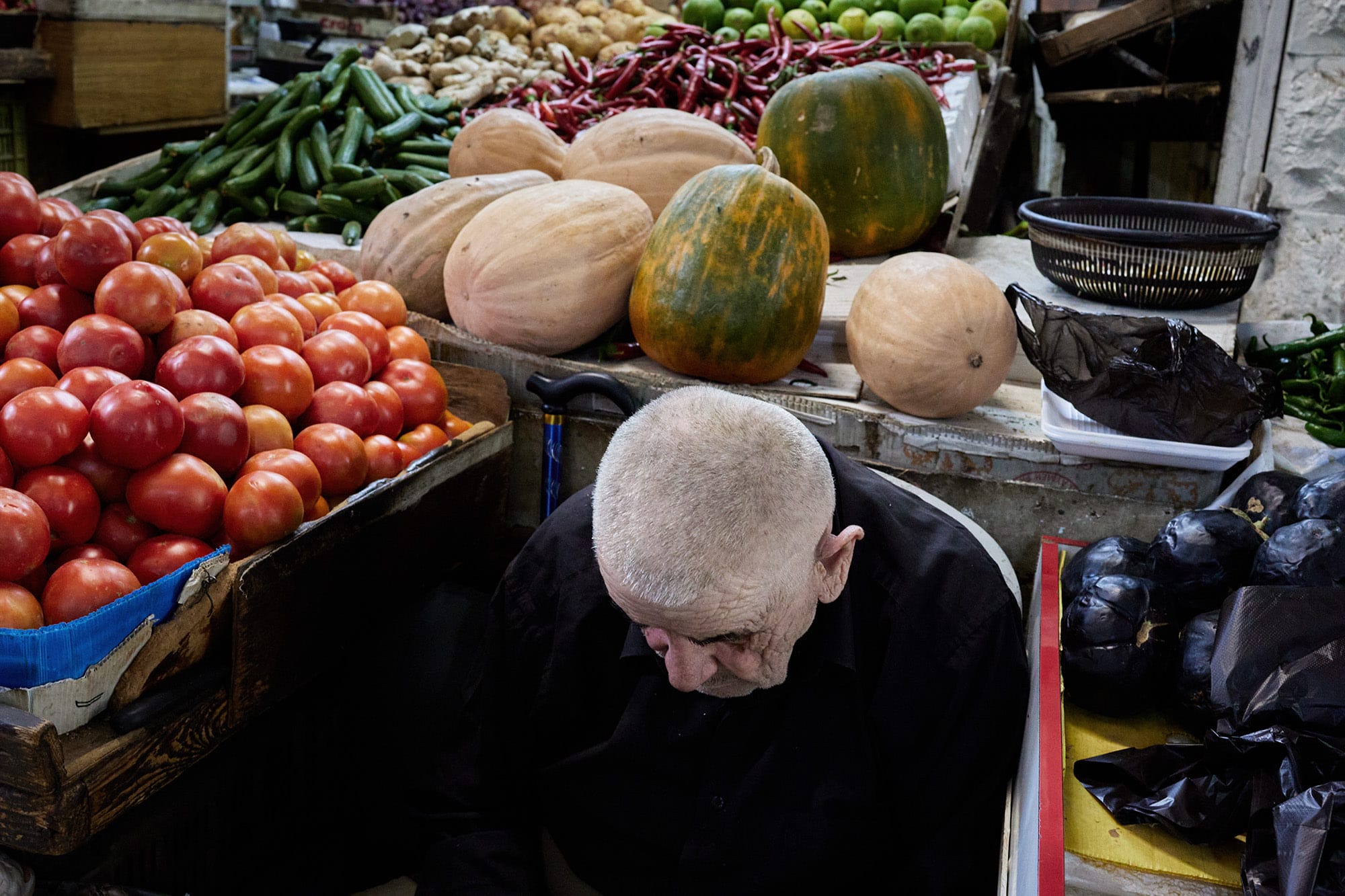 A candid street photograph by Janusz Jurek of a man seated in his market stall, with melons next to his head