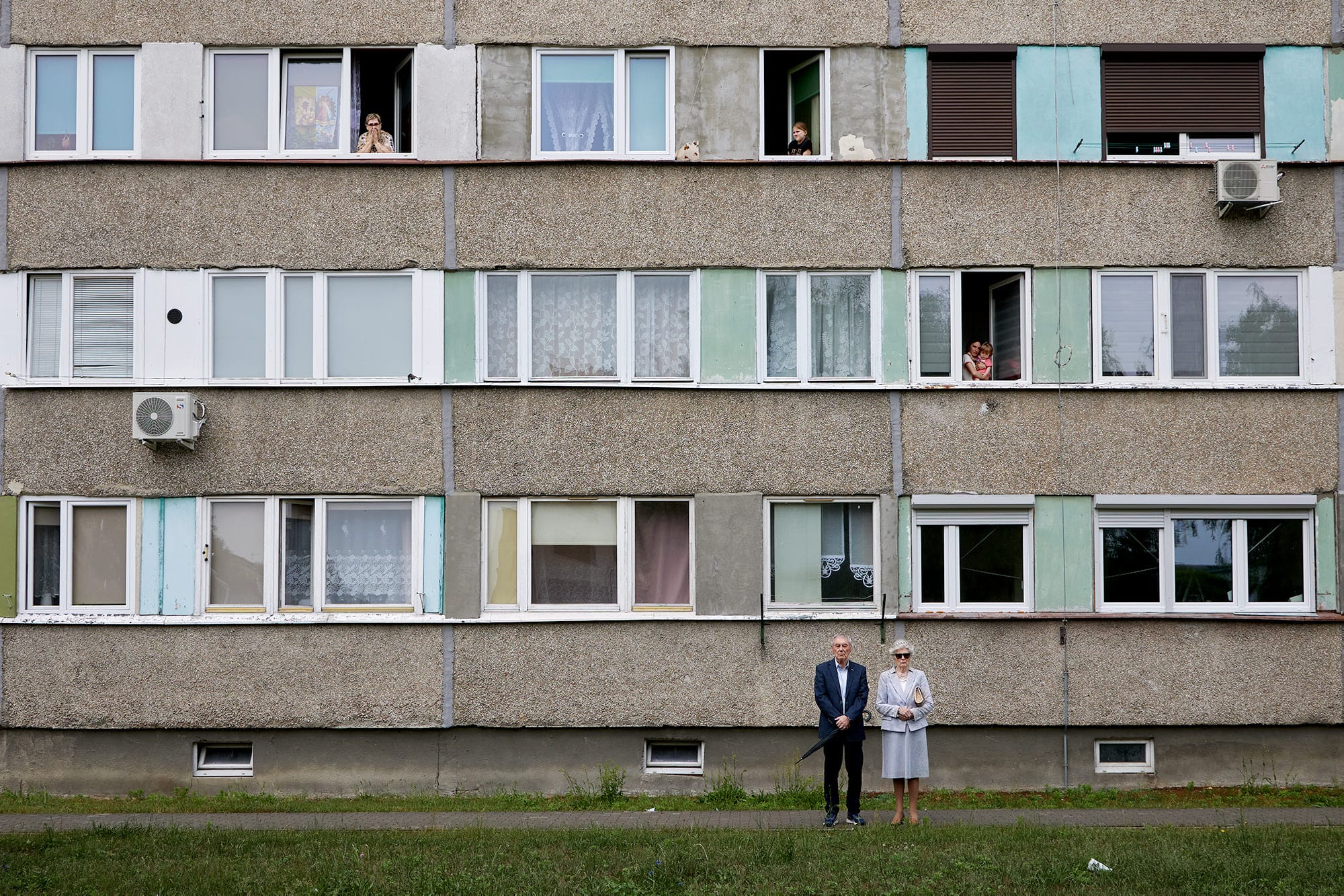 A candid street photograph by Janusz Jurek of an older couple standing in front of an apartment building, with other people observing from their windows