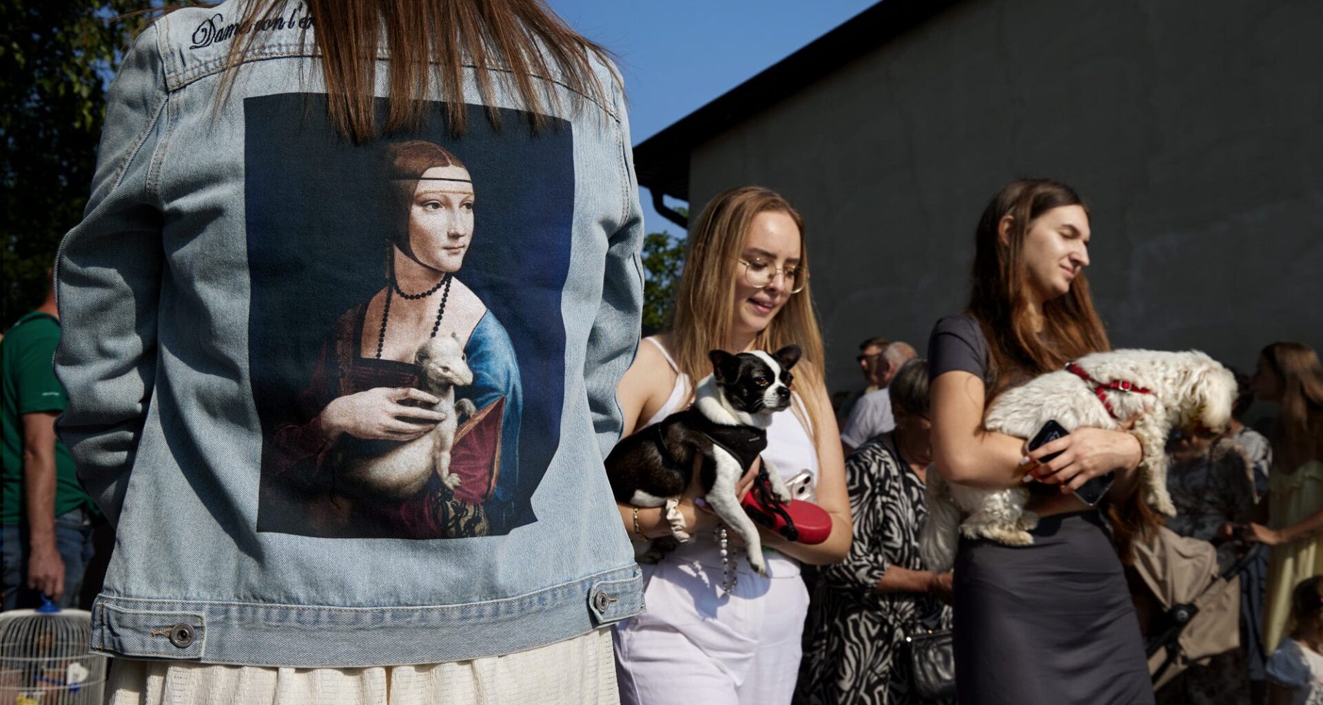 A candid street photograph by Janusz Jurek of women carrying dogs, with a denim jacket with a Leonardo Da Vinci painting of a woman holding an ermine