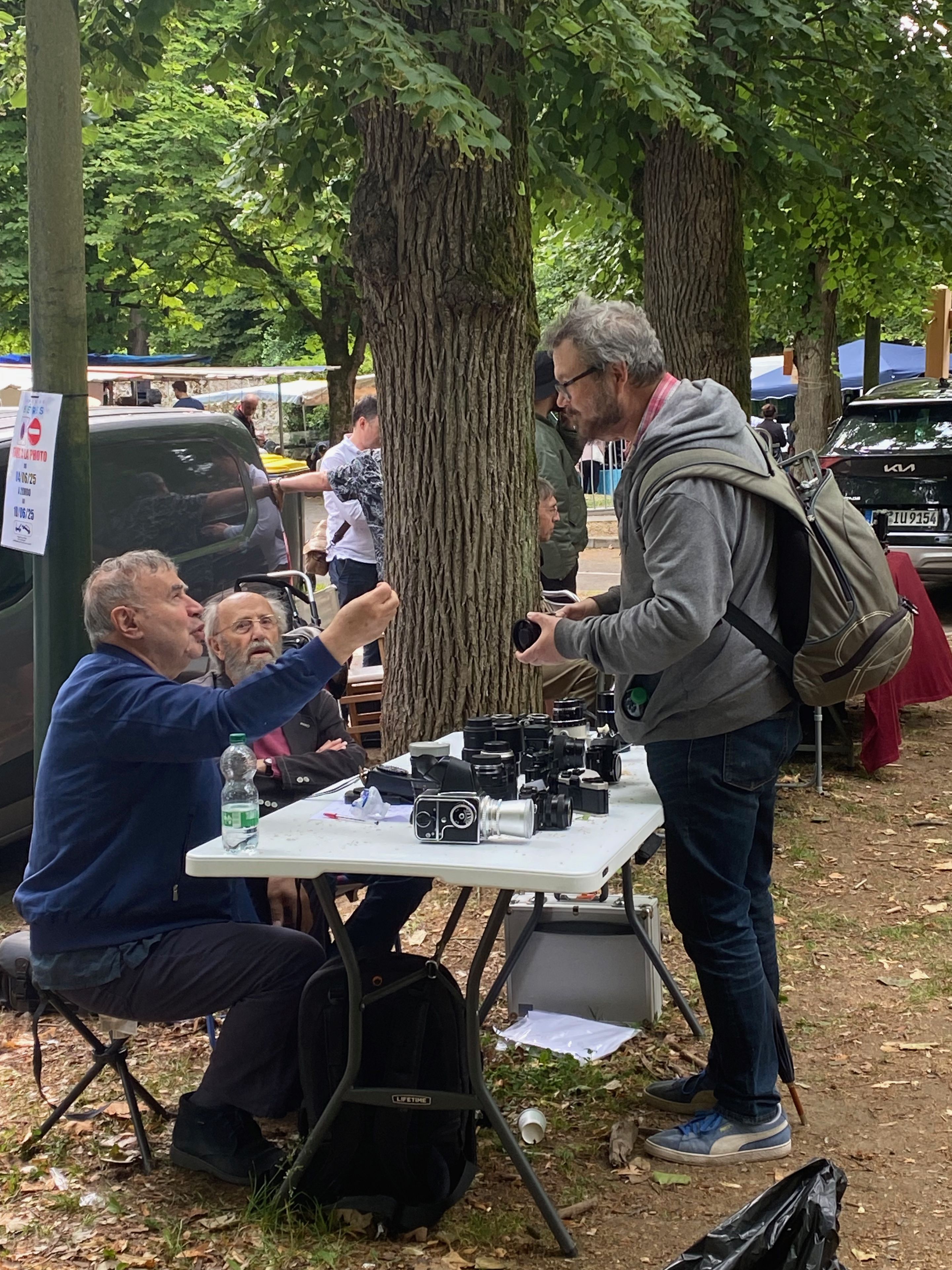 A seated vendor gestures animatedly while discussing a camera with a standing visitor, a camera and several lenses laid out on the table between them.