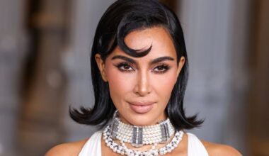 A woman with shoulder-length dark hair styled in soft waves wears a pearl choker and a white outfit, posing against a blurred background. Her makeup is glamorous with defined brows and nude lips.