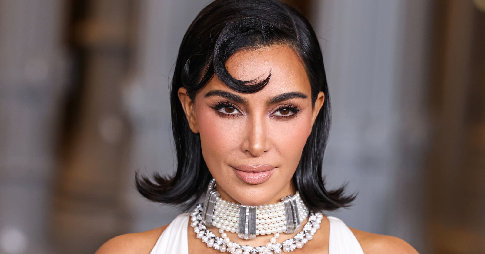A woman with shoulder-length dark hair styled in soft waves wears a pearl choker and a white outfit, posing against a blurred background. Her makeup is glamorous with defined brows and nude lips.